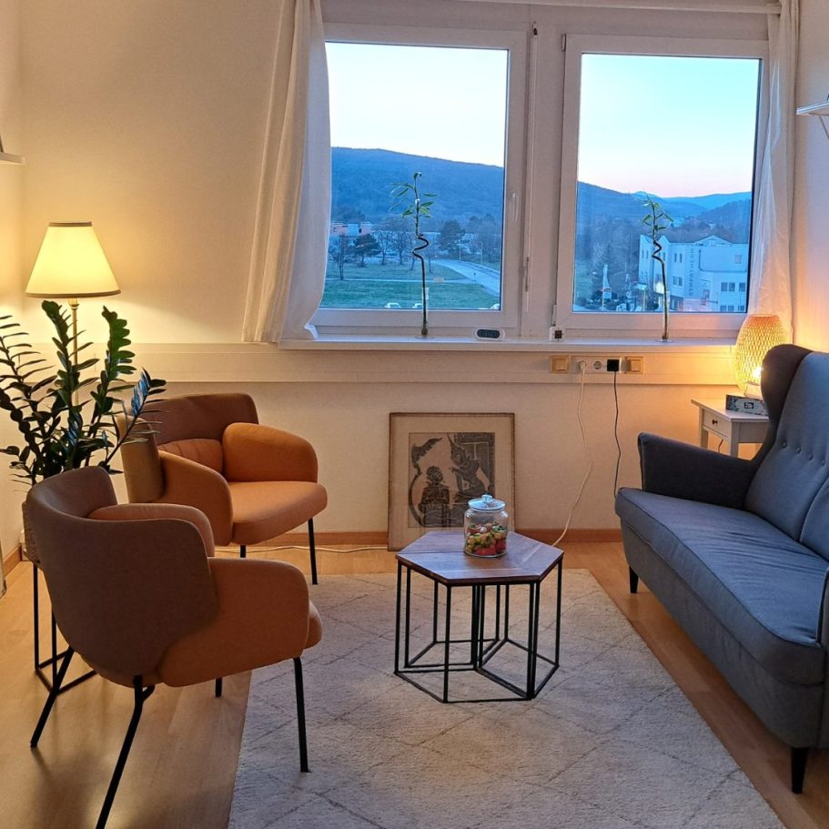 Helle Wohnzimmerszene mit Sofa, Sesseln, Tisch und Blick auf die Landschaft.