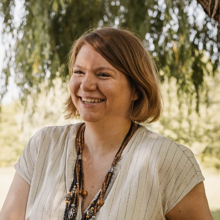 Portrait von Nadja Daunicht: Lächelnde Frau mit kurzen blonden Haaren, im Freien vor grünen Bäumen.