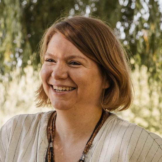 Portrait von Nadja Daunicht: Lachende Frau mit kurzem, braunem Haar und heller Kleidung, vor grünem Hintergrund.